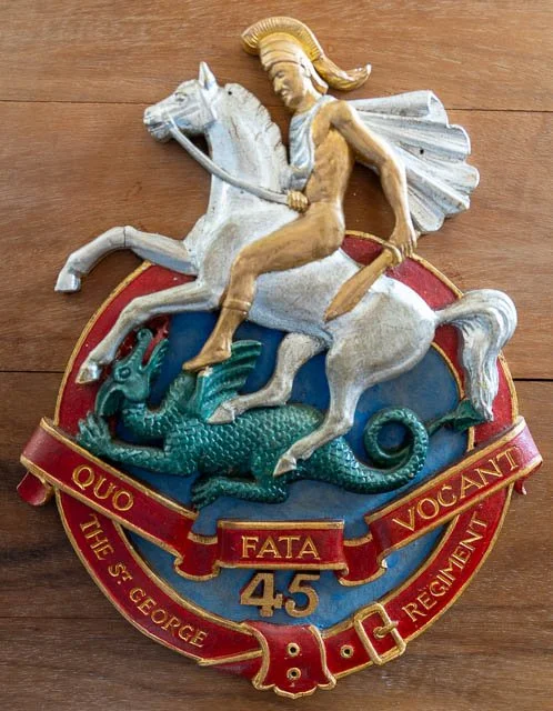 A decorative emblem of the 45th Infantry Regiment, also known as the 'Fighting 45s,' featuring a soldier riding a white horse and fighting a dragon, with a red banner containing golden text.