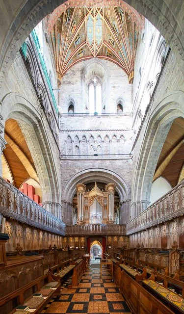Interior of a historic church or cathedral with high vaulted ceilings, ornate colorful decorations on the ceiling, stone walls, and an organ in the background, with wooden pews on either side of a patterned floor.