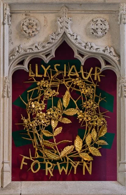 A decorative memorial plaque with the name Llyswau Forwyn, featuring a gold wreath of leaves and flowers on a purple background within an ornate white stone frame.