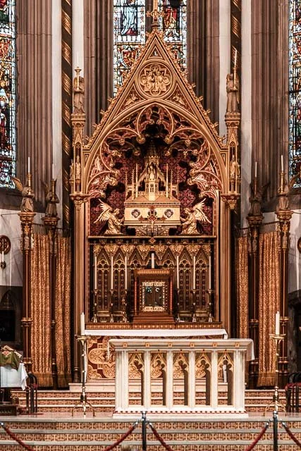 Interior of a Gothic-style church altar with intricate gold and wood details, statues, and stained glass windows.