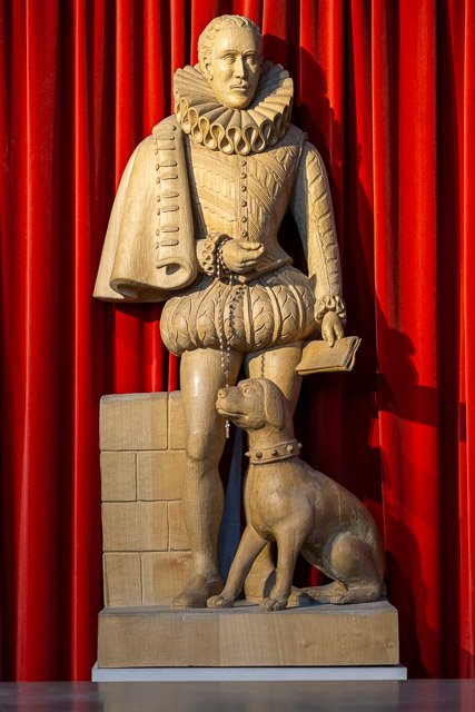 Stone statue of William Shakespeare dressed in Elizabethan clothing, holding a scroll and book, with a dog sitting beside him, against a red curtain background.