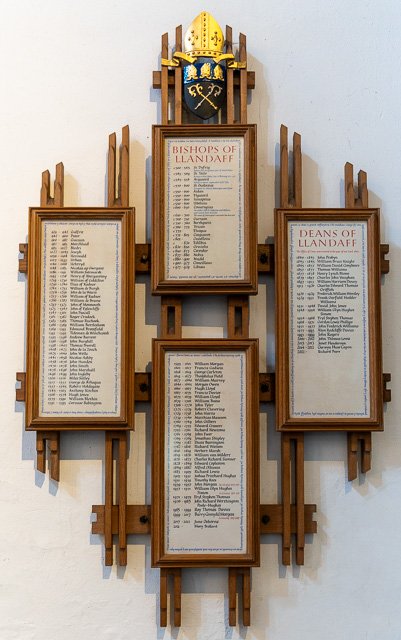 Wooden wall display with framed lists of names, labeled as Bishops of Llandaff and Deans of Llandaff, and a crest featuring a helmet and shield at the top.