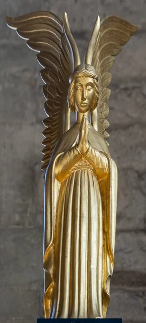 Gold-colored statue of a woman with wings, hands in prayer, eyes closed, standing on a base against a gray background.