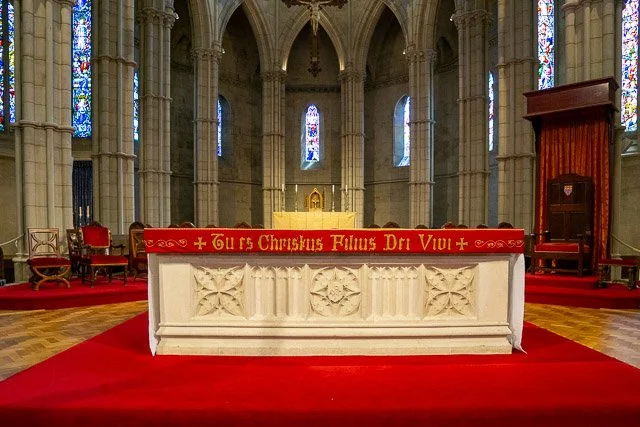 Interior of a church with an altar covered in a white cloth, surrounded by a red carpet, with stained glass windows in the background.