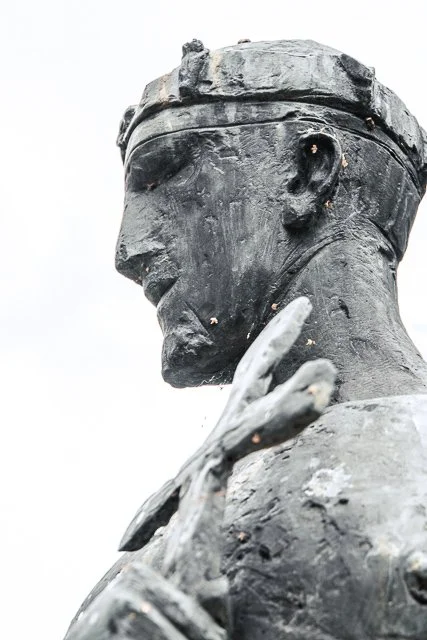 A close-up side profile of a weathered bronze statue of a person with glasses, holding a hand near the face.