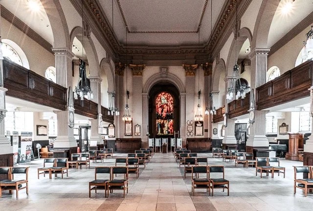 Interior of a church or cathedral with rows of chairs, arched windows, and a stained glass window at the front.