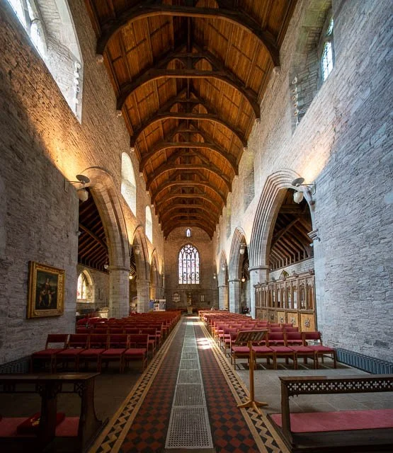 Interior of a church with wooden vaulted ceiling, stone walls, stained glass windows, rows of red chairs, and religious artwork.