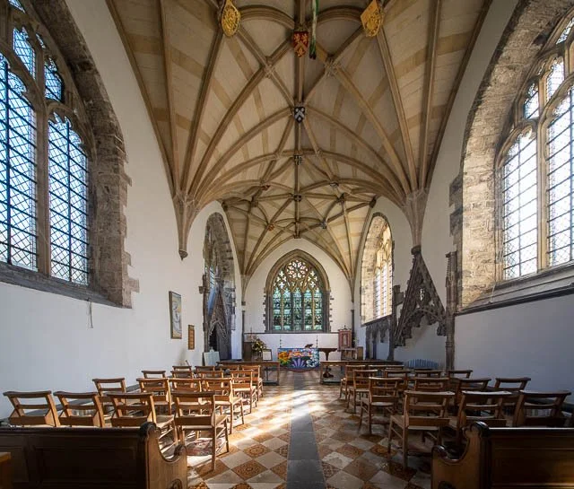Interior of a historic chapel with high vaulted ceiling, stained glass windows, wooden pews, and an altar at the front decorated with flowers.