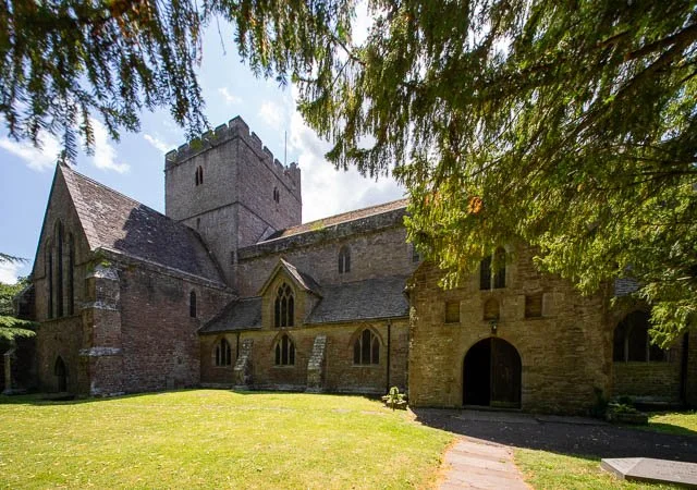An old stone church with a tower, surrounded by a green lawn and large trees, under a partly cloudy sky.