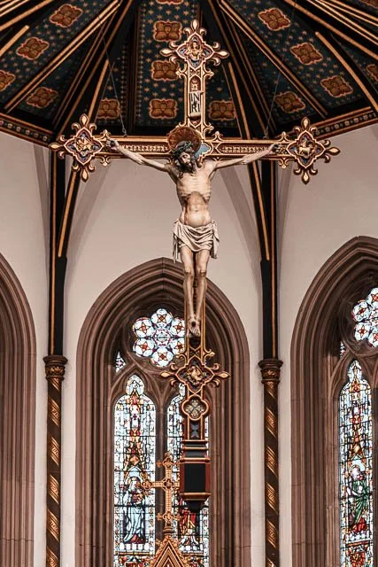 Crucifix hanging inside a church with stained glass windows behind it.