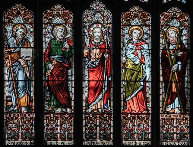 Colorful stained glass window depicting five religious figures, each with a halo, holding objects, with decorative floral patterns at the bottom.