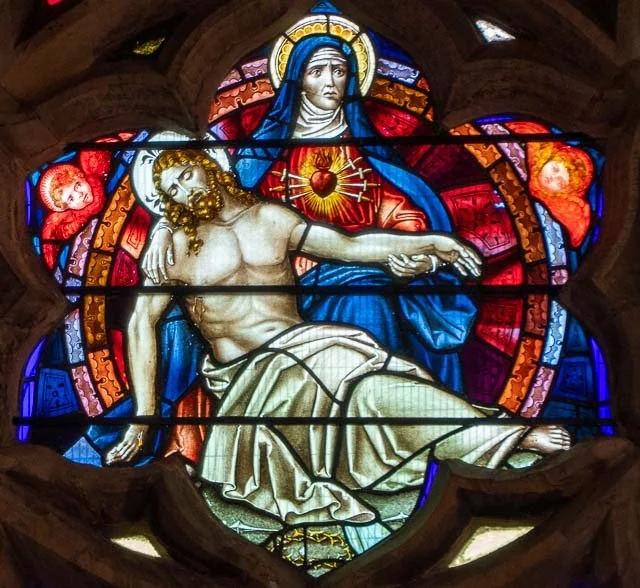 Stained glass window depicting the body of Jesus Christ after crucifixion, with a halo around his head, set in a religious building.