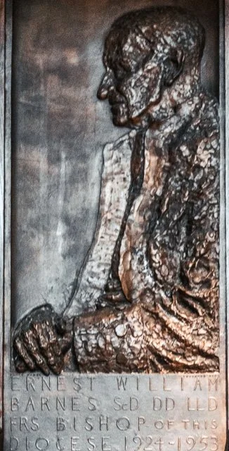 Bronze statue of a man in profile, sitting with head bowed and hands resting on his lap, mounted on a stone base with engraved name and dates.