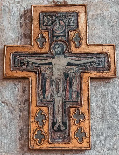 Painted wooden crucifix depicting Jesus Christ on the cross, with figures and symbols around him, mounted on a textured gray wall.