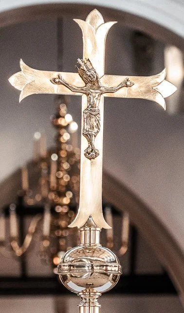 A silver crucifix with Jesus Christ on the cross, mounted on an ornate base, in front of a blurred chandelier in a church setting.
