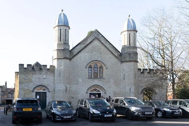 A historic church with two small towers and a larger central arch, surrounded by parked cars and leafless trees.