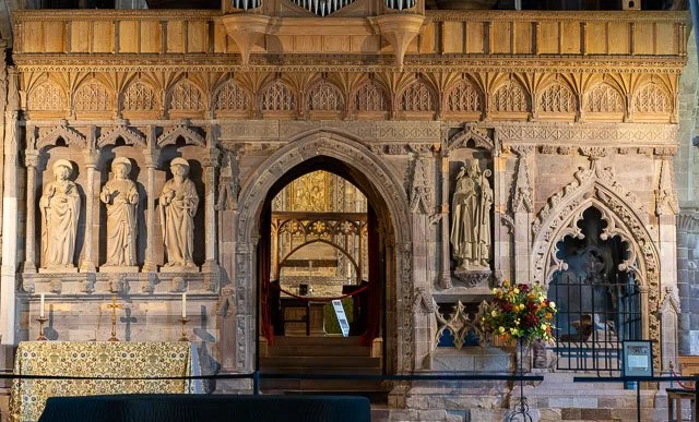 Stone wall with carved statues and decorative arches, and an arched doorway leading to an interior with a wooden rail and a display with flowers outside.
