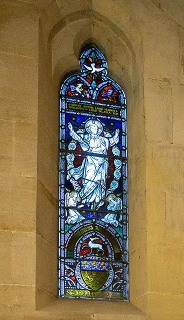 A stained glass window in a church depicting a religious figure, possibly Jesus Christ, with arms raised and surrounded by angels and decorative elements.