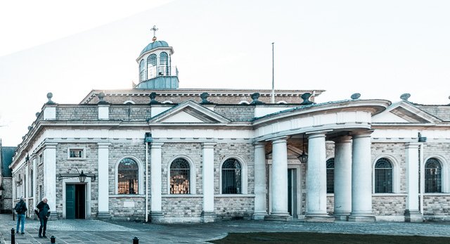 Exterior of a historic stone building with columns, arched windows, and a small dome on top, with people walking nearby.