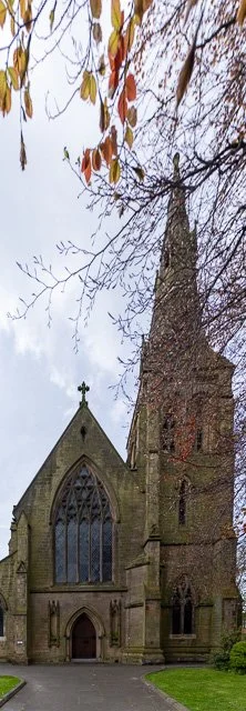 View of a historic stone church with gothic architectural features, including tall pointed windows and a steeple, surrounded by trees with some autumn leaves.
