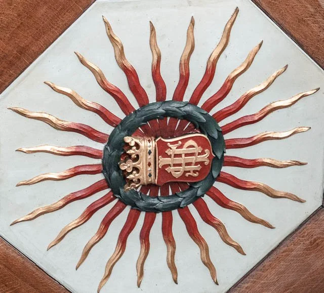 Decorative wall art featuring a central red shield with a gold crown on top, surrounded by a black wreath and radiating gold and red flame-like spikes.