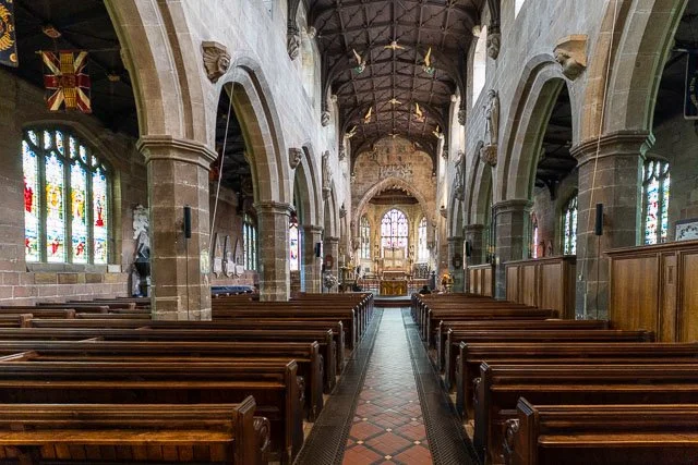 Interior of a church with wooden pews on either side of a central aisle, stone arches, stained glass windows, and an altar at the front.