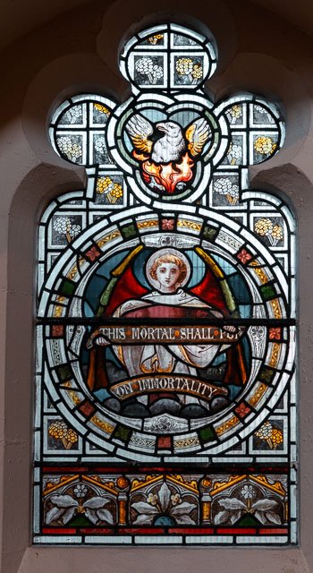 Stained glass window featuring an angel in the center holding a banner that reads 'This mortal shall put on immortality,' with a dove and colorful decorative glass above.