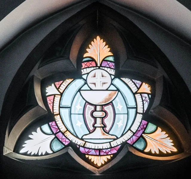 Colorful stained glass window featuring a chalice with a cross and a host, decorated with leaves and geometric patterns.