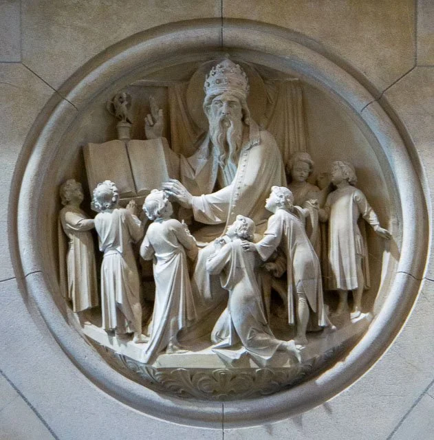 Stone relief sculpture depicting a biblical scene with a central figure wearing a crown and robes, surrounded by eight people in various poses, some listening and others gesturing, set within a rounded arch.
