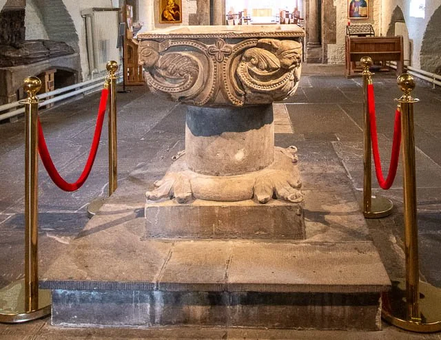 Ancient stone baptismal font with carved ornamental design, surrounded by red velvet ropes in a church or cathedral.