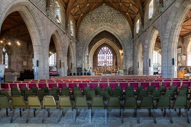 Interior of a church with stone walls and arched ceilings, rows of green and maroon chairs facing an altar with a stained glass window.