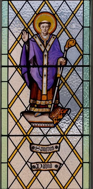 Stained glass window depicting a saint with a halo, dressed in purple and gold robes, holding a staff in one hand and raising the other in blessing, with a scroll bearing the word 'Sanctus' below.