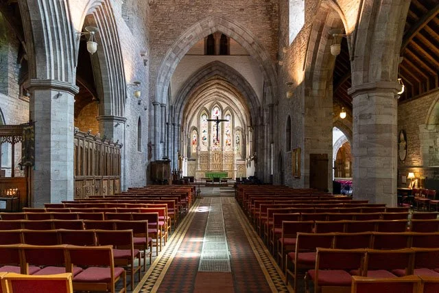 Interior of a church with rows of wooden pews leading to an altar featuring a large cross and stained glass windows.