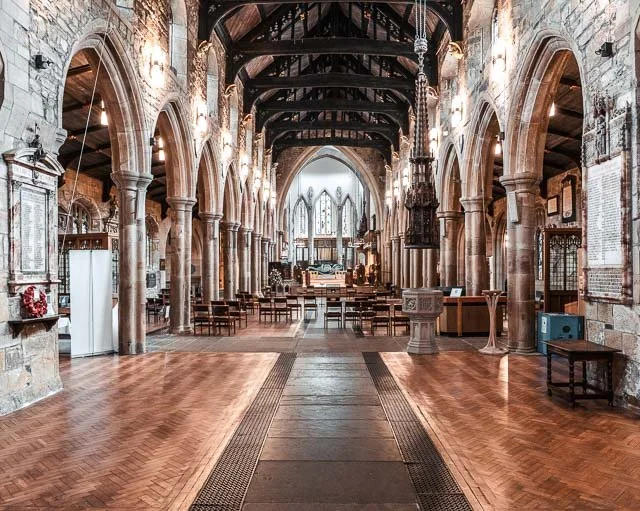 The interior of a historic church with stone walls, wooden arches, and a high vaulted ceiling. Wooden pews are arranged in rows facing the altar at the front, with stained glass windows behind it lighting the space.