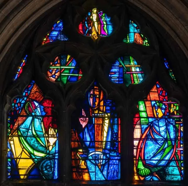 Colorful stained glass window with religious imagery, featuring figures and intricate designs, in a Gothic cathedral style.