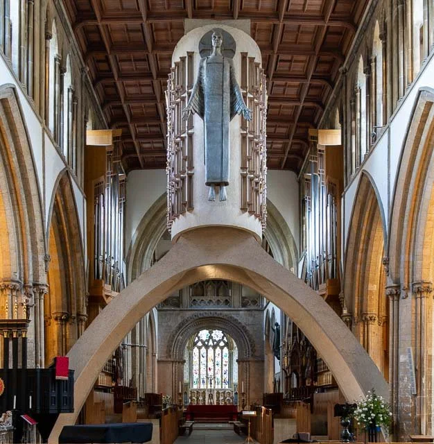 Interior of a church featuring a large baptismal font with a sculpture of a saint or religious figure above it, stained glass windows, and wooden architectural details.