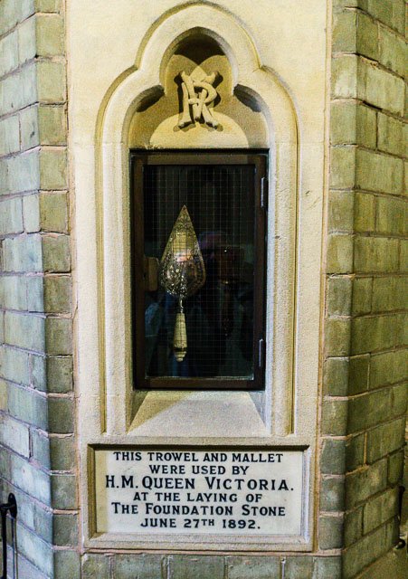 A small niche with a scroll-shaped arch, containing a decorative droplet-shaped object with a tassel, above a plaque indicating it was used by Queen Victoria during her laying of the foundation stone on June 27, 1892.