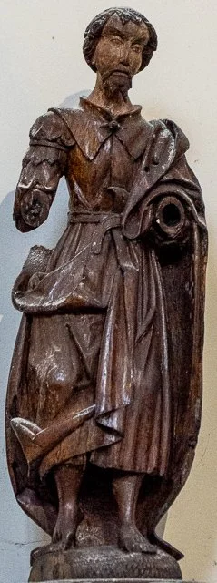 Wooden statue of a woman in historical dress, holding a spool of thread, with a serious expression.
