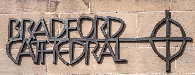 Metal sign reading 'Bradford Cathedral' with a circular cross symbol mounted on a brick wall.