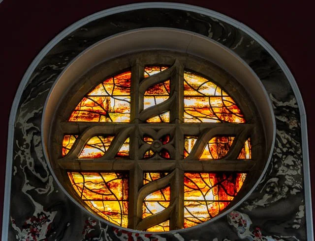 A stained glass window with shades of orange, yellow, and red, featuring a decorative cross design, viewed through a circular opening with surroundings of dark marble or stone.