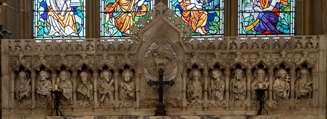 Interior of a church with a row of stone statues of saints and biblical figures beneath colorful stained glass windows.