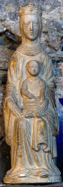 Wooden sculpture of a crowned woman holding a smaller figure, set against a stone background.