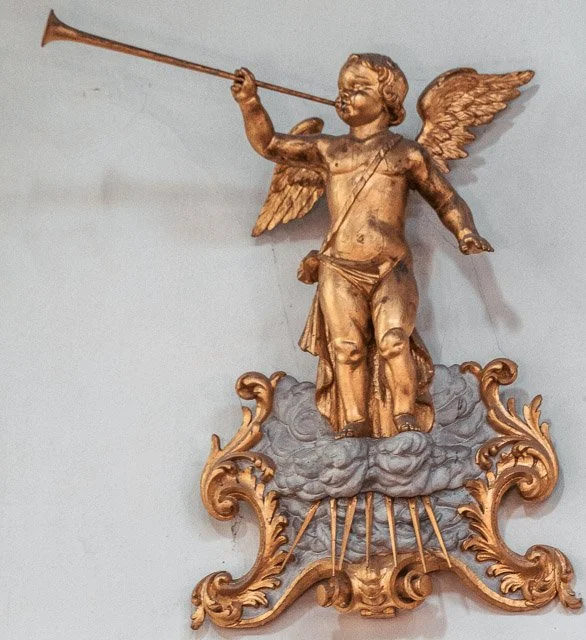 A gold-colored sculpture of a cherub with wings, holding a trumpet, standing on a cloud with decorative scrolls.