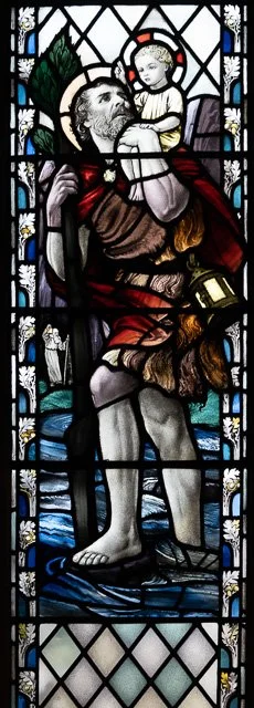 Stained glass window depicting a man carrying a child on his shoulder, with a serene expression. The scene includes a blue background and floral details around the figures.