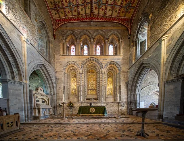 Interior of a historic church with stone walls, stained glass windows, and an ornate altar decorated with gold accents and religious artwork.