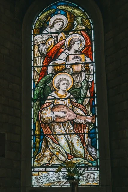 Stained glass window depicting Jesus Christ, angel musicians, and a woman with a halo playing a guitar, in a church setting.