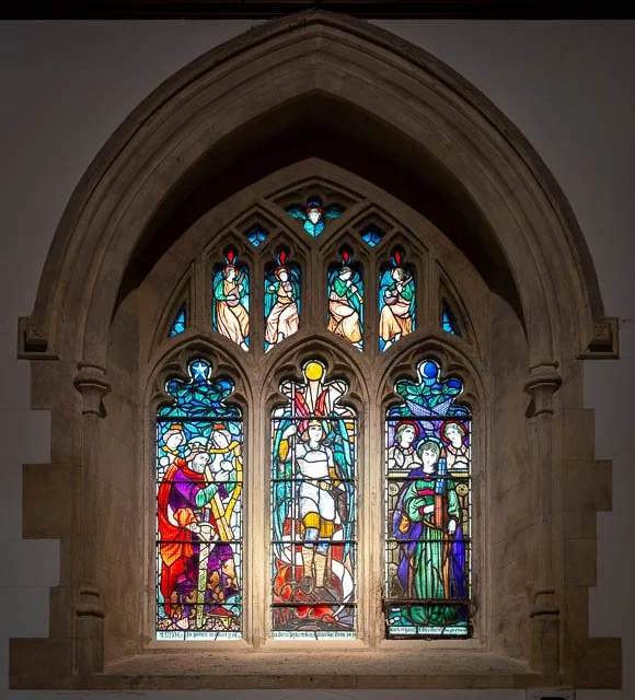 Colorful stained glass window depicting religious figures and scenes in a Gothic-style stone frame.