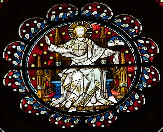 Stained glass window depicting Jesus Christ seated on a throne with a halo, holding a scepter and a globe, surrounded by ornate architecture and decorative floral patterns.