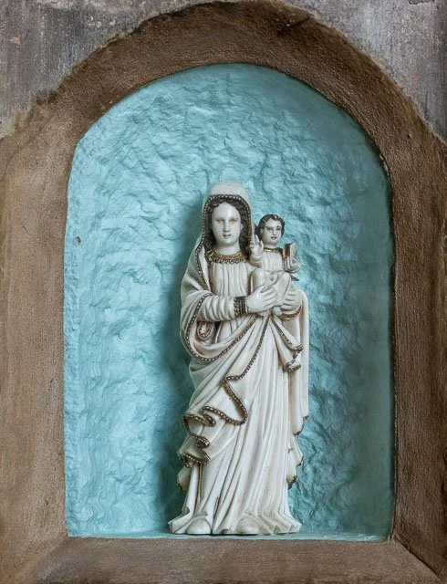 Statue of the Virgin Mary holding baby Jesus, set in a small arched niche with a textured blue background and brown border.
