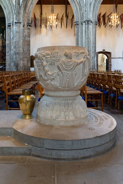 A stone fountain with a carved sculpture of two figures on a circular pedestal inside a church or cathedral, with rows of wooden chairs and hanging banners in the background.